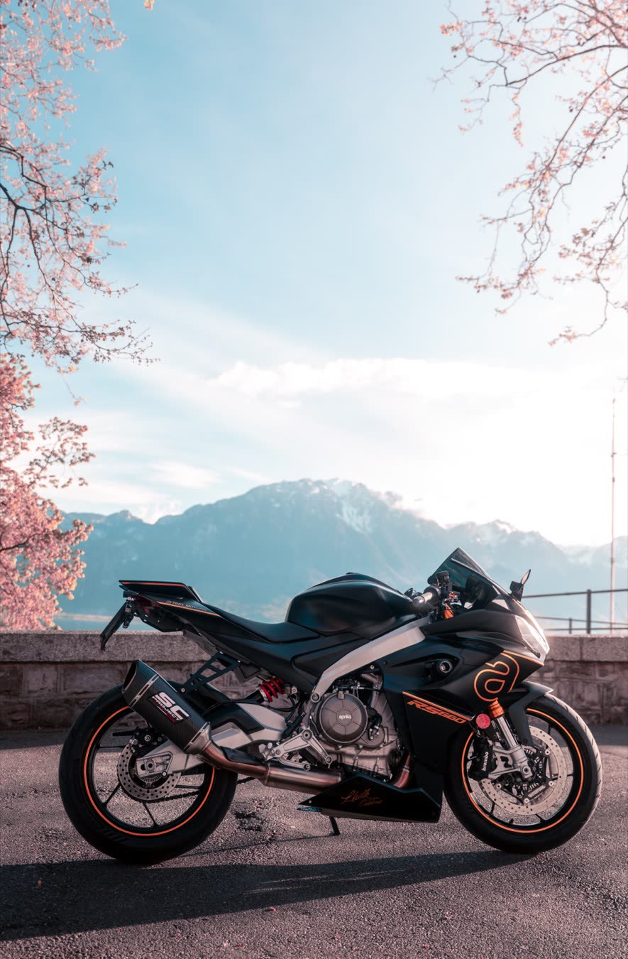 Moto sportive noire et orange à l'arrêt devant un panorama de montagnes, cerisiers en fleurs au premier plan.