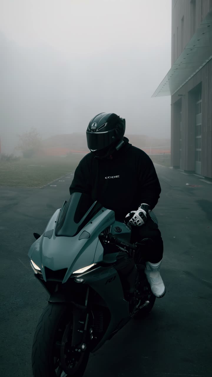 Motard à l'arrêt sur sa sportive dans le brouillard, devant un bâtiment industriel.